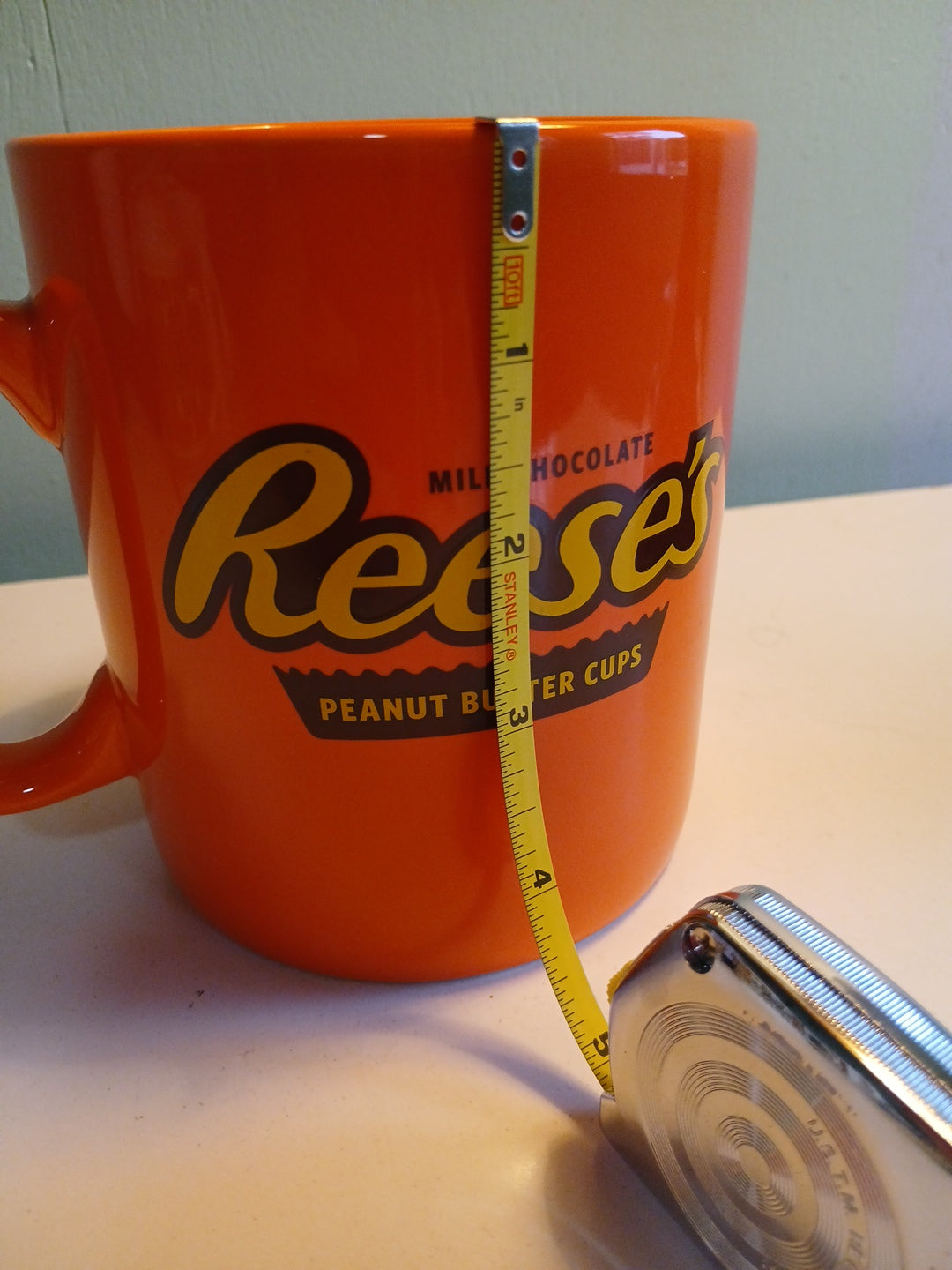 Orange mug with Reese's logo being measured with a tape measure on a light surface.