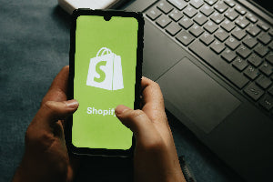 Person holding a smartphone with Shopify app on a desk with laptop and mouse