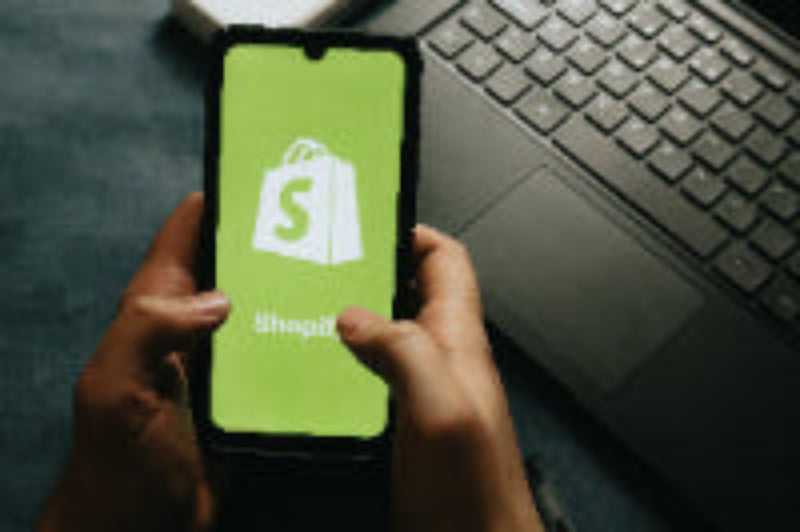 Person holding a smartphone with an app icon displayed, next to a laptop on a desk.