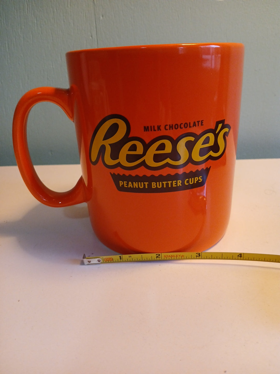 Orange mug with Reese's logo next to a measuring tape on a light-colored wall.